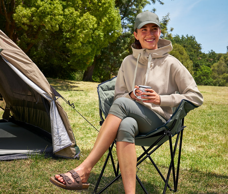 Une femme assise dans une chaise de camping Tambu »ASANA« tient une tasse et porte un legging 3/4 et un sweat à capuche.