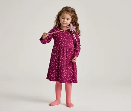 Une fillette aux cheveux bouclés tient une baguette magique, porte une robe bordeaux à motifs floraux et des collants roses.