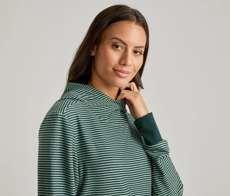 Une femme touche son cou et sourit, portant un sweat à capuche rayé vert et blanc avec des poignets foncés.
