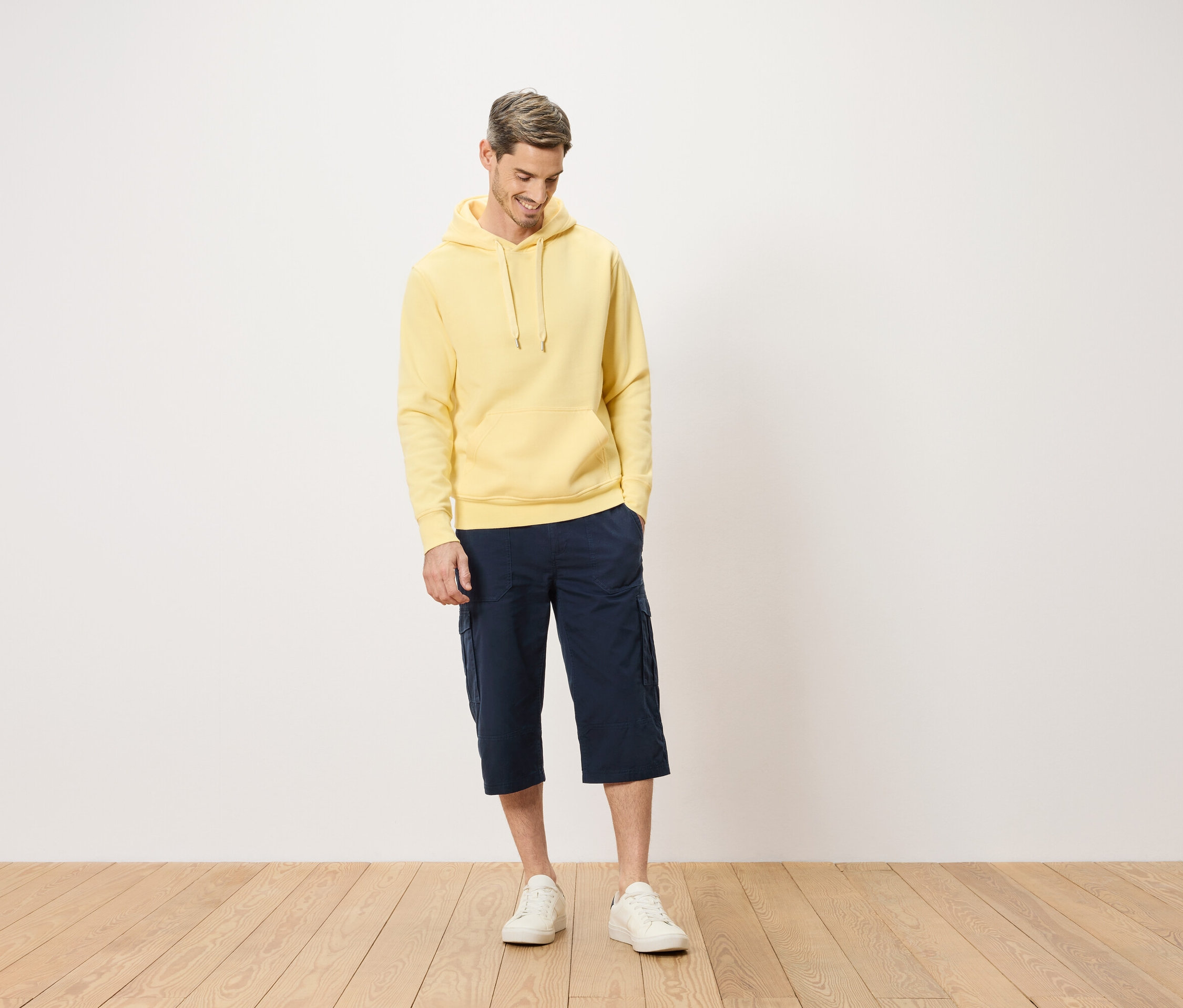 Homme portant un sweat à capuche jaune et un short cargo bleu debout sur un plancher en bois.