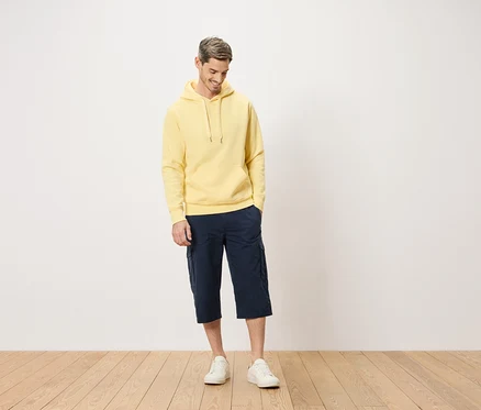 Homme portant un sweat à capuche jaune et un short cargo bleu debout sur un plancher en bois.