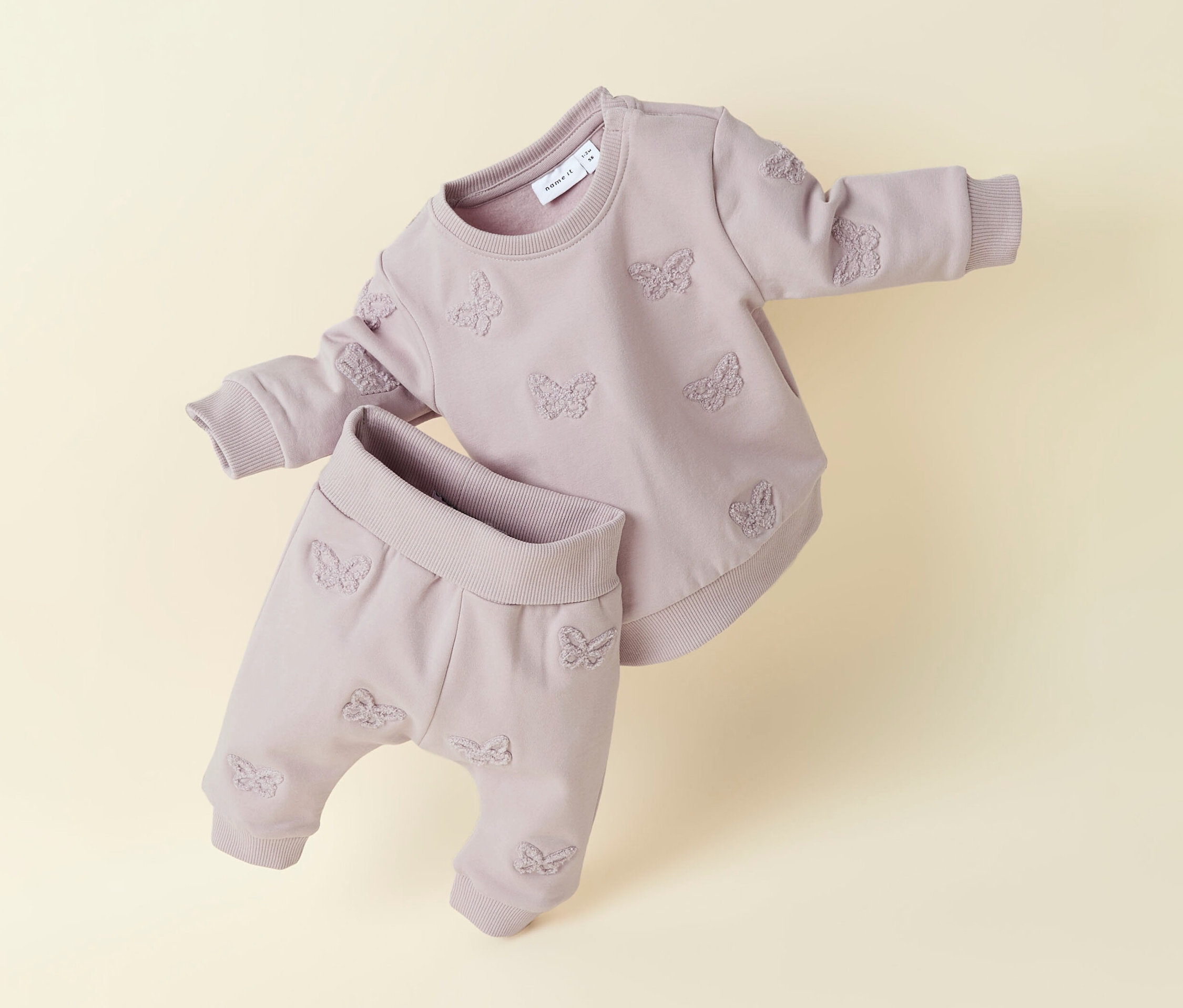 Un sweat-shirt pour bébé NAME IT et un pantalon assorti lilas avec un motif de papillons sont posés sur un fond beige.