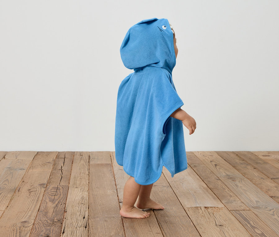 Un enfant se tient pieds nus sur un plancher en bois et porte un poncho bleu à capuche.