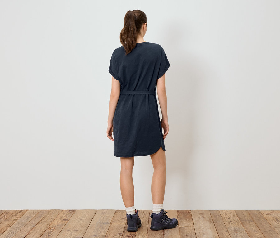 Vue arrière d'une femme portant une robe t-shirt bleu marine ceinturée et des chaussures de randonnée devant un mur blanc sur un plancher en bois.