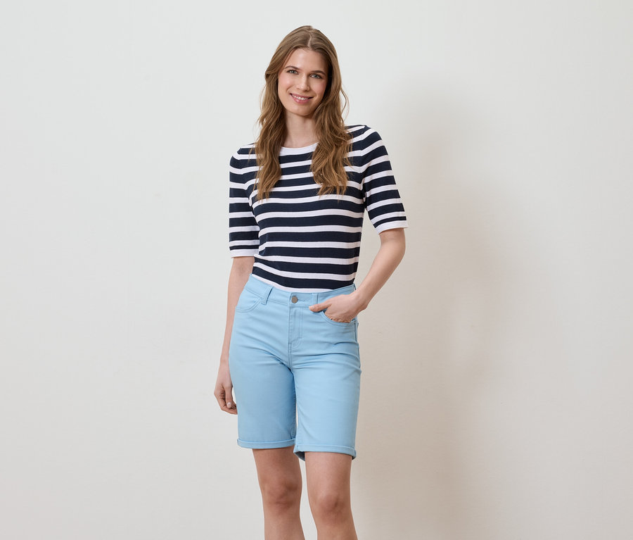 Une femme se tient devant un fond clair, elle porte un t-shirt rayé et un short bleu clair.