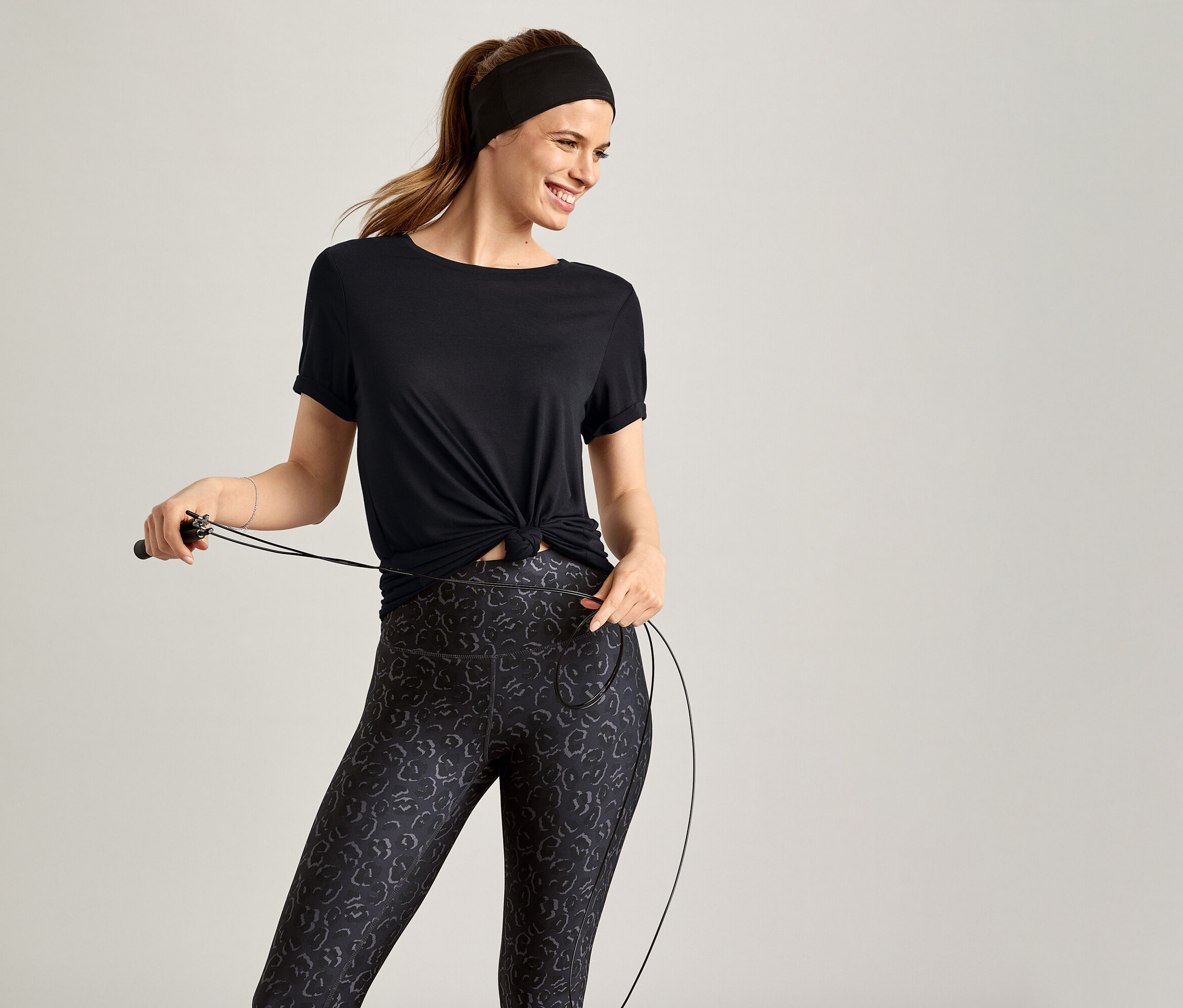 Femme portant un T-shirt de sport noir tenant une corde à sauter sur fond gris.