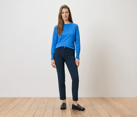 Une femme porte un chemisier bleu et un pantalon bleu marine avec des mocassins noirs. Elle se tient sur un plancher de bois devant un mur blanc.
