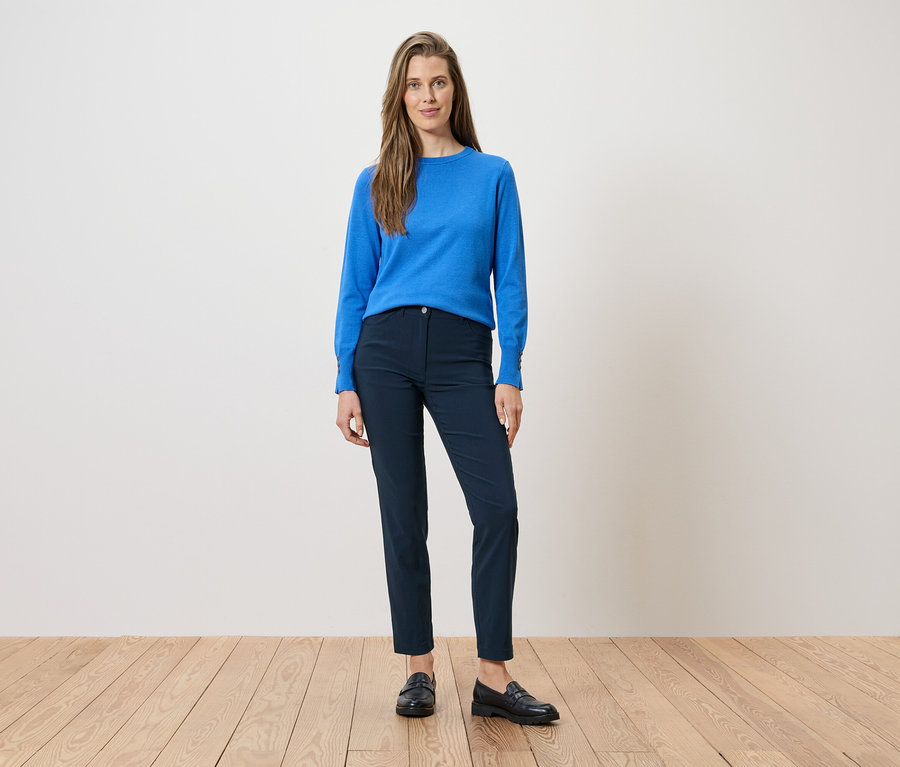 Une femme porte un chemisier bleu et un pantalon bleu marine avec des mocassins noirs. Elle se tient sur un plancher de bois devant un mur blanc.
