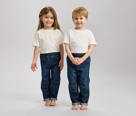 Deux enfants, un blond et un brun, se tiennent pieds nus côte à côte, portant des t-shirts blancs et des jeans bleus avec des motifs d'ours.
