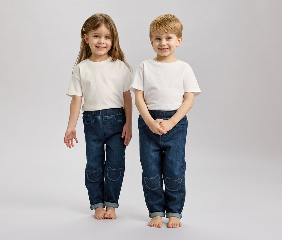 Deux enfants, un blond et un brun, se tiennent pieds nus côte à côte, portant des t-shirts blancs et des jeans bleus avec des motifs d'ours.