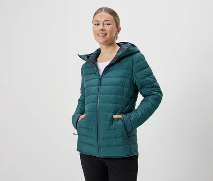 Une femme souriante portant une veste matelassée à capuche émeraude et un pantalon noir sur un fond neutre.