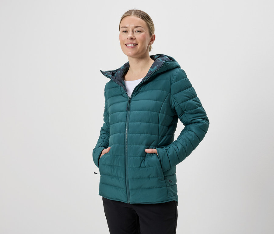 Une femme souriante portant une veste matelassée à capuche émeraude et un pantalon noir sur un fond neutre.