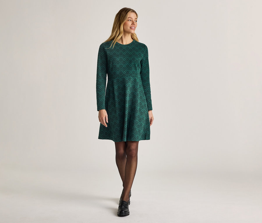 Femme portant une robe verte à motifs à manches longues, des collants et des chaussures sur un fond clair.