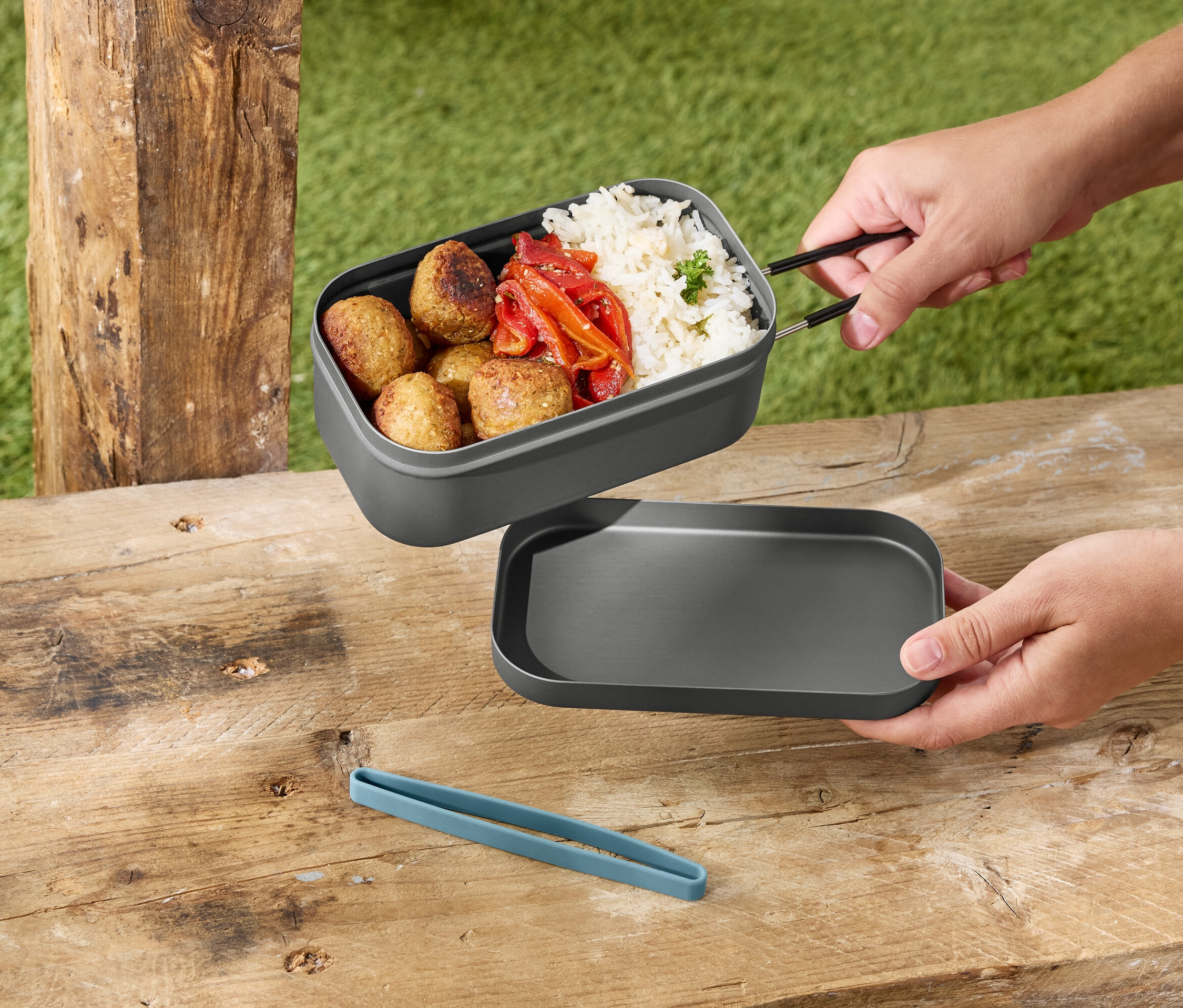 Des mains tiennent une boîte à lunch grise contenant du riz, des boulettes de viande et des poivrons au-dessus d'un couvercle gris sur une table en bois.
