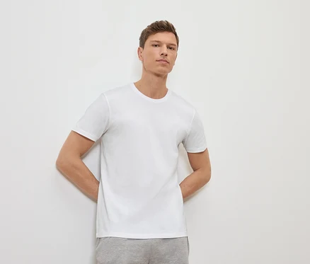 Un homme appuyé contre un mur blanc portant un t-shirt blanc et un pantalon de survêtement gris.