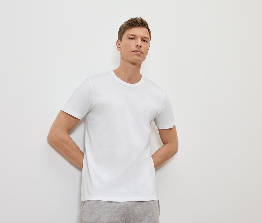 Un homme appuyé contre un mur blanc portant un t-shirt blanc et un pantalon de survêtement gris.