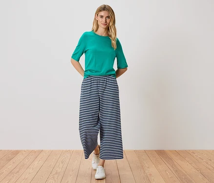 Femme debout, mains dans les poches, portant un t-shirt vert, un pantalon rayé bleu et blanc et des chaussures blanches.