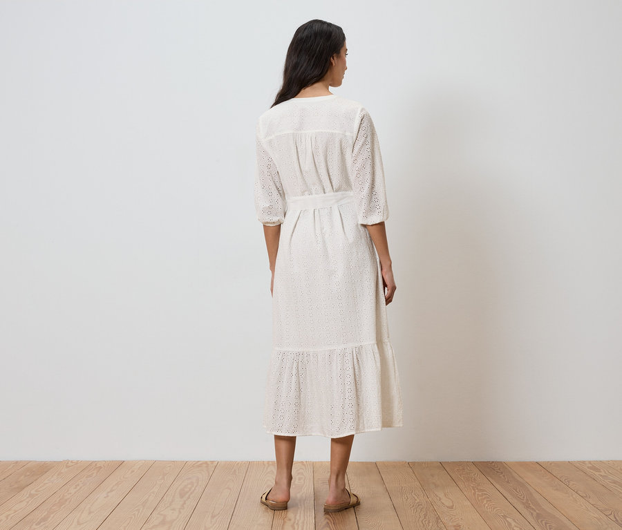 Une femme se tient dos à la caméra, portant une robe blanche avec une ceinture et des détails ajourés.
