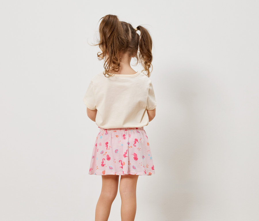Vue arrière d'une petite fille avec des tresses, portant un t-shirt crème et une jupe rose avec des motifs marins.