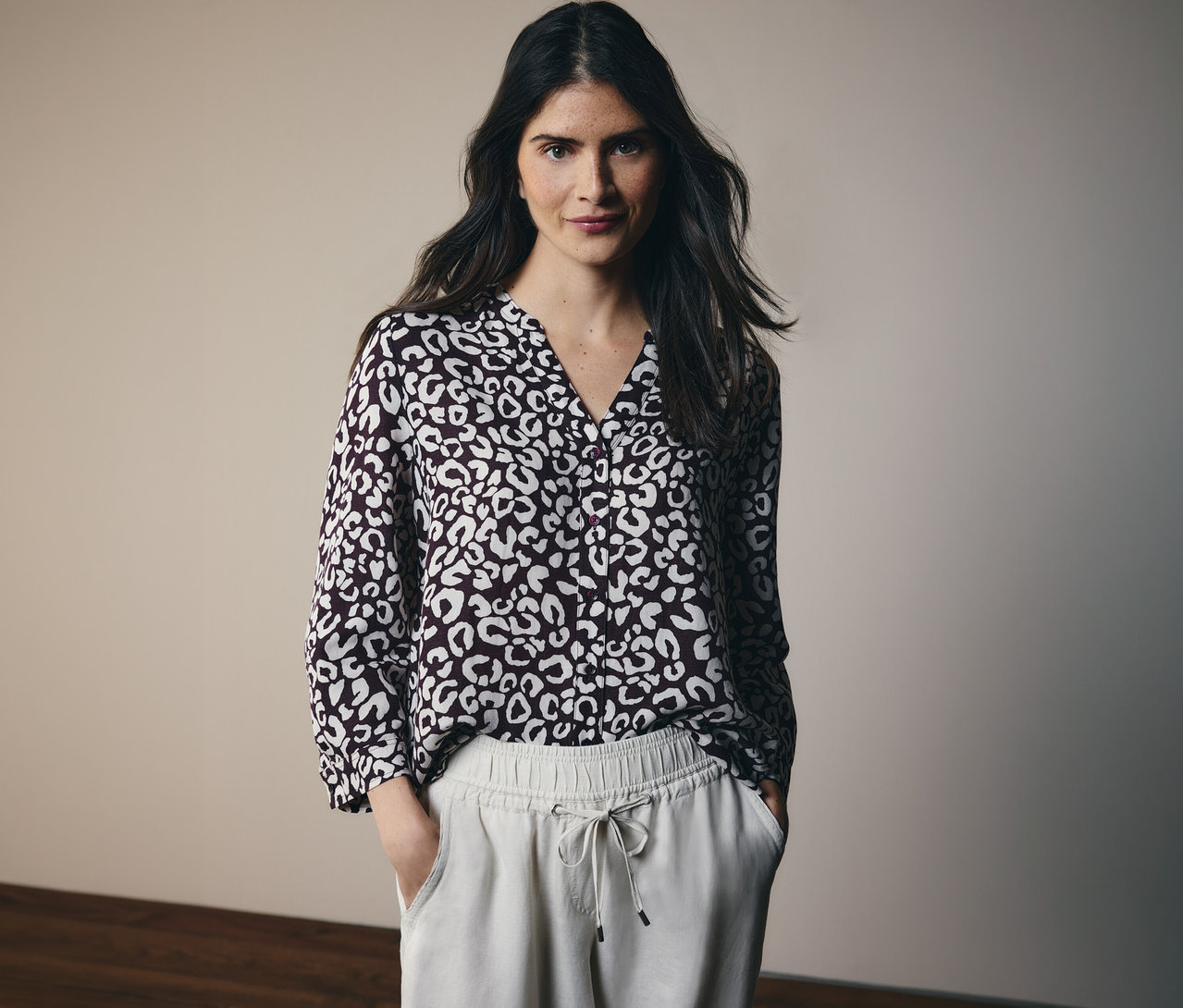 Une femme aux longs cheveux foncés porte un chemisier à motifs et un pantalon clair à cordon.