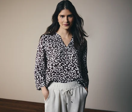 Une femme aux longs cheveux foncés porte un chemisier à motifs et un pantalon clair à cordon.