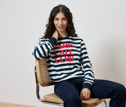 Une femme est assise sur une chaise et porte un sweat-shirt rayé avec l'inscription "Paris Cafe Club" et un jean.
