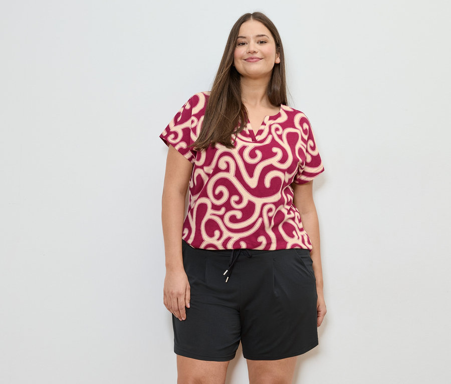 Une femme aux cheveux bruns porte un chemisier à motifs rouge et beige et un short noir sur fond blanc.