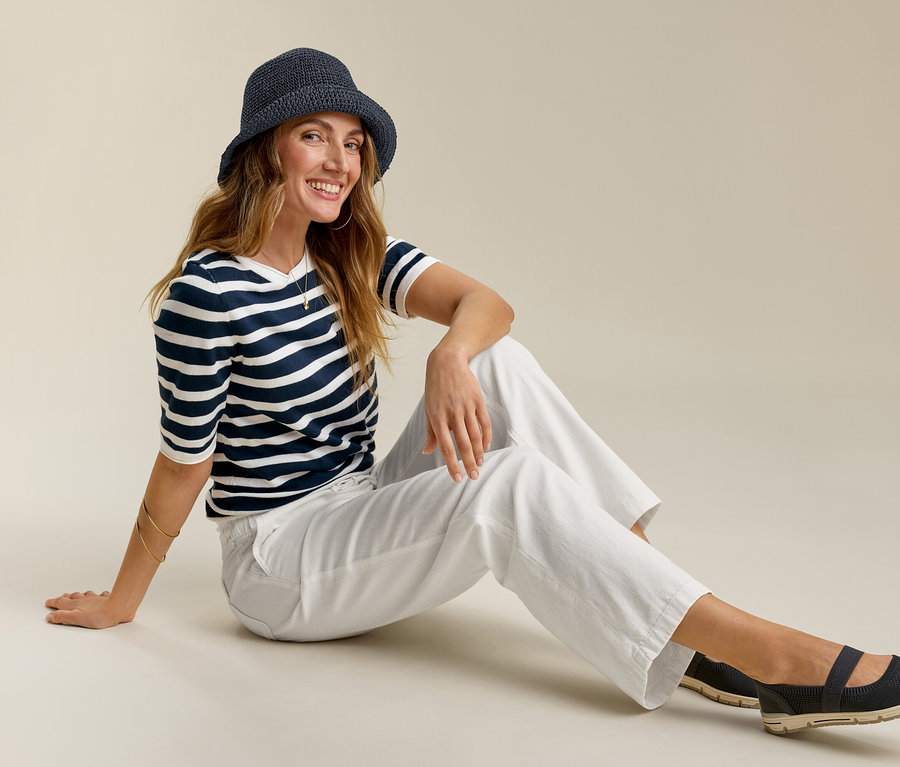 Femme assise portant un jeans grand confort, un t-shirt rayé et un chapeau. Elle sourit.