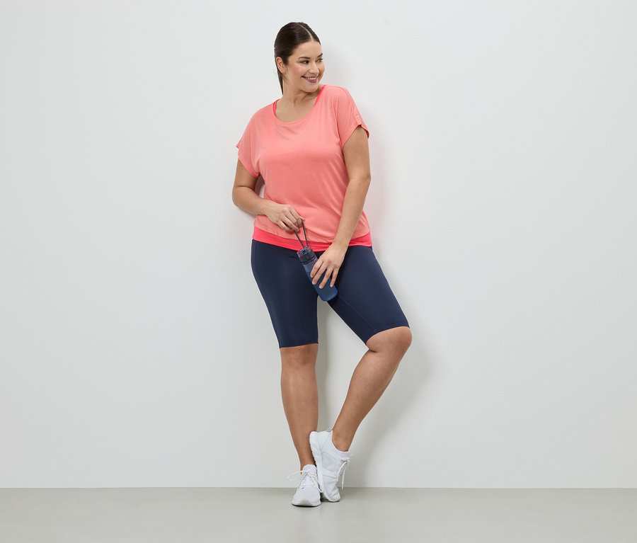 Une femme en tenue de sport avec une bouteille d'eau est appuyée contre un mur. Elle porte un t-shirt corail, un short bleu et des baskets blanches.