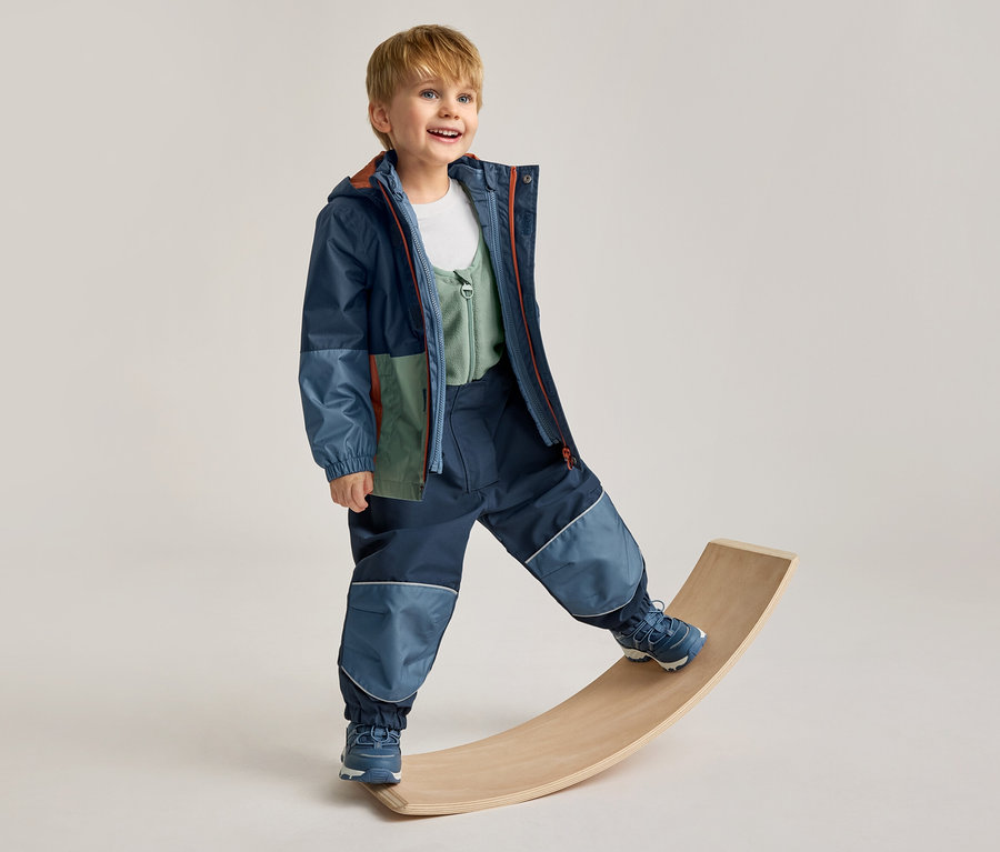 Garçon souriant portant une veste multicolore et un pantalon bleu foncé avec des empiècements bleu clair, en équilibre sur une planche en bois.