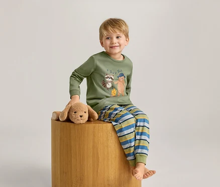 Un garçon blond est assis sur un tabouret à côté d'un chien en peluche. Il porte un pyjama rayé vert olive.