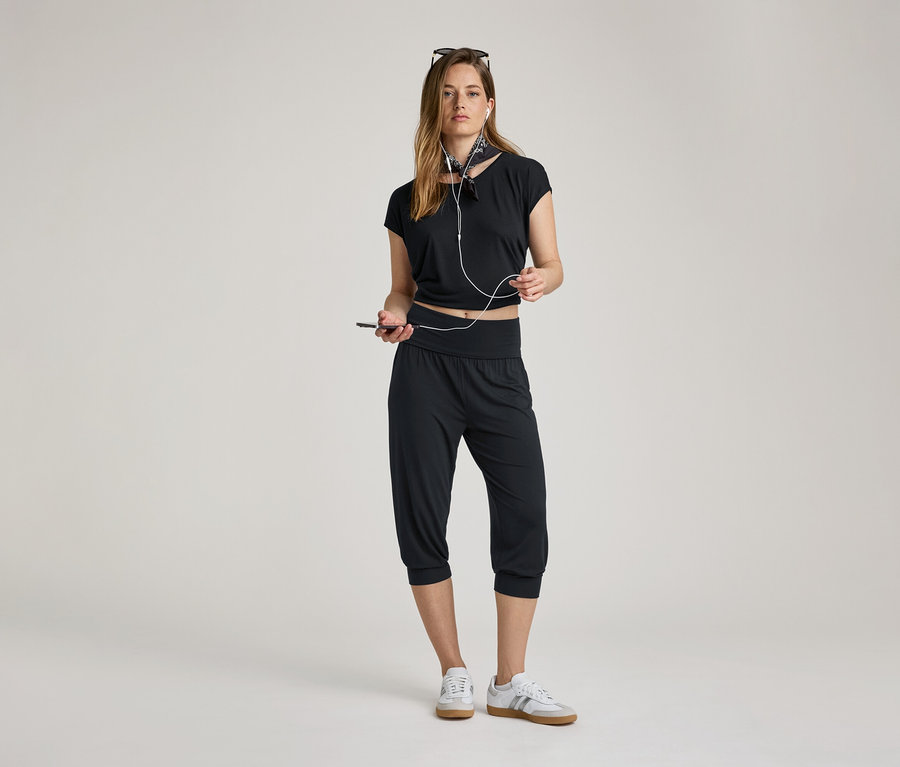 Femme avec des écouteurs et un téléphone. Elle porte un pantalon 3/4 de yoga, un haut noir et des baskets.