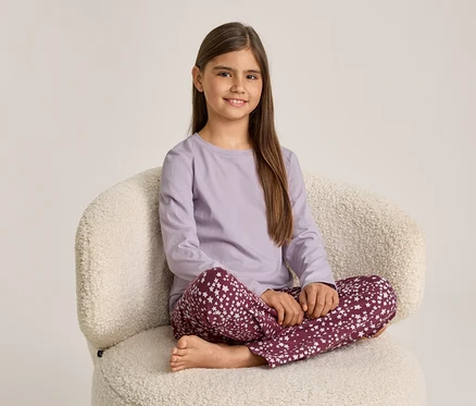 Une fille souriante est assise en tailleur sur un fauteuil crème. Elle porte un haut à manches longues violet clair et un pantalon rouge foncé à motifs d'étoiles blanches.