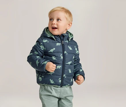 Un jeune garçon blond porte une veste matelassée bleu marine à motifs de dinosaures et un pantalon vert.