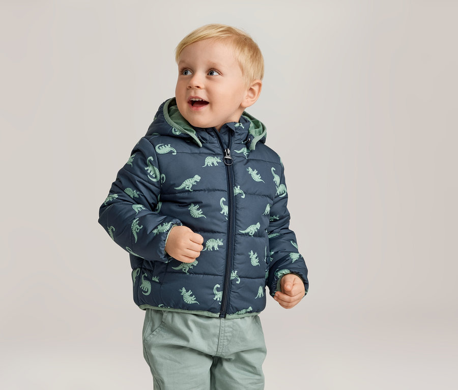 Un jeune garçon blond porte une veste matelassée bleu marine à motifs de dinosaures et un pantalon vert.