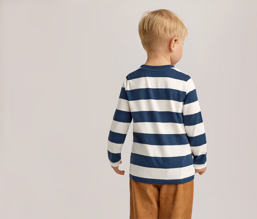 Vue arrière d'un enfant blond portant un t-shirt à manches longues rayé bleu et blanc et un pantalon en velours côtelé marron sur un fond clair.