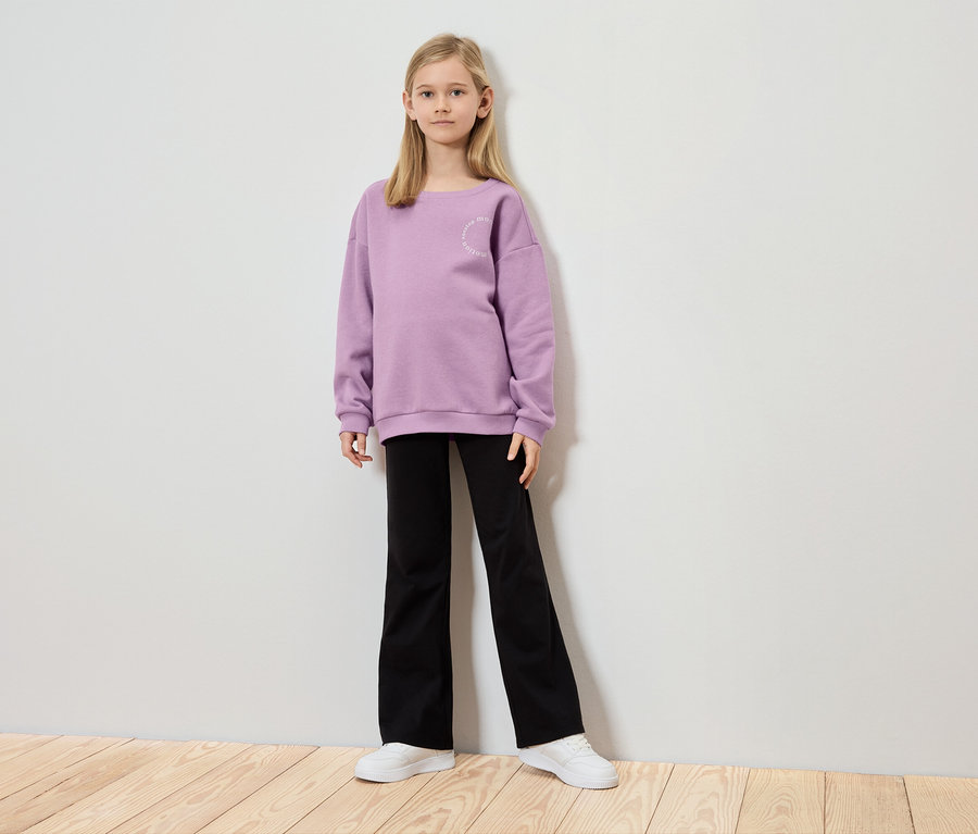 Une fille se tient devant un mur, portant un pull violet, un pantalon noir et des chaussures blanches.