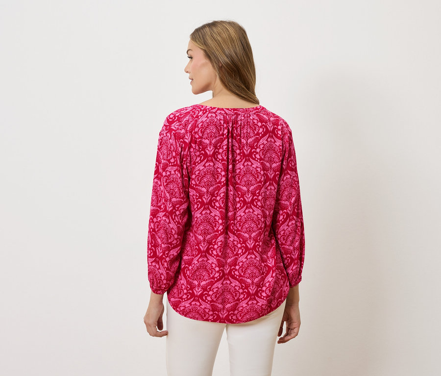 Vue de dos d'une femme aux cheveux blonds portant un chemisier rose à manches longues avec des motifs et un pantalon blanc.