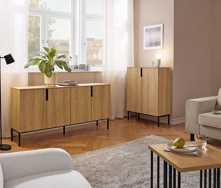 Un salon présentant un long buffet en bois clair sous une fenêtre et une armoire haute assortie. Un lampadaire noir, deux fauteuils, un tapis shaggy et une table basse avec une poire et un verre d'eau sont également visibles.