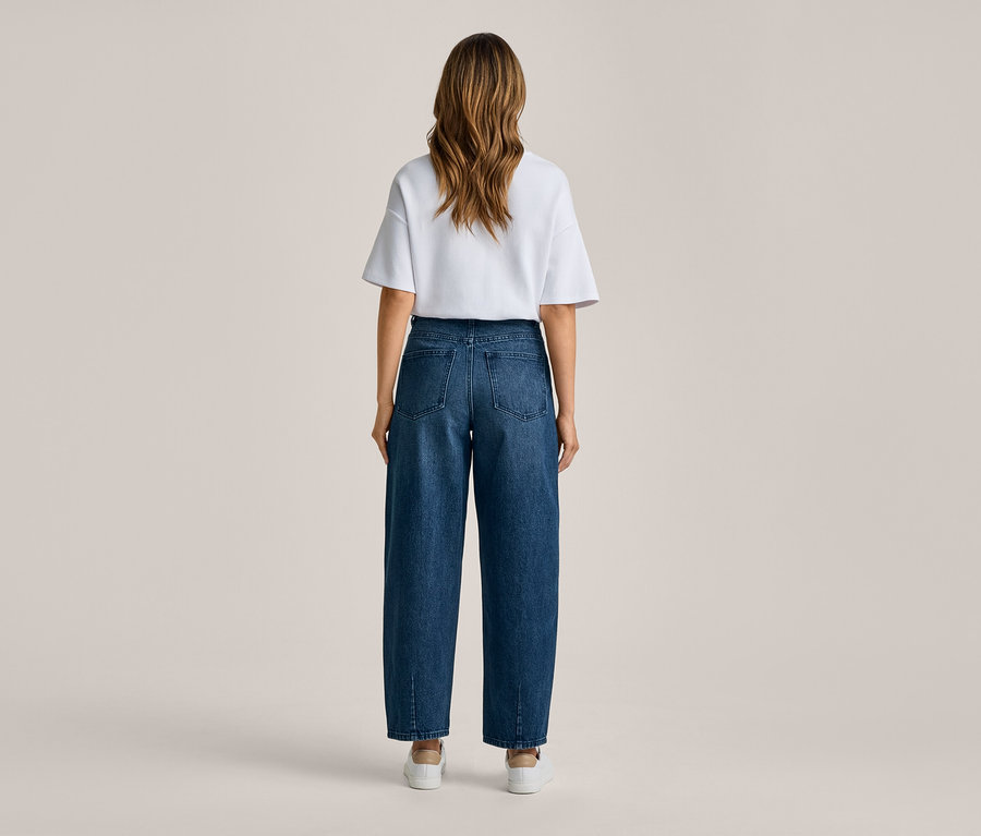 Femme debout de dos, portant un t-shirt blanc, un jean large bleu foncé et des baskets blanches.