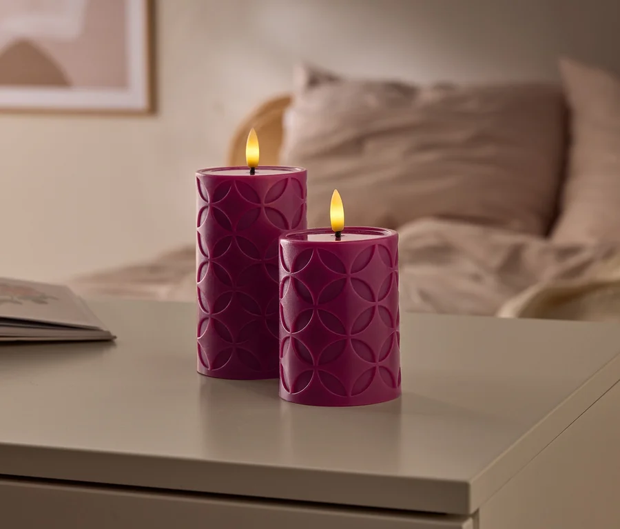 Deux bougies LED violettes à motifs géométriques sont allumées sur une table, à côté d'un livre ouvert, avec un lit flou en arrière-plan.