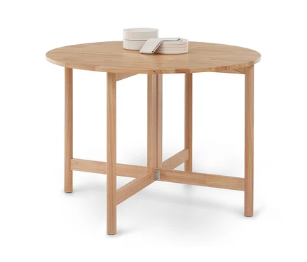 Table ronde en bois clair à quatre pieds et entretoises, surmontée de deux bols blancs empilés, d'une pile de petites assiettes blanches et d'une serviette claire pliée.