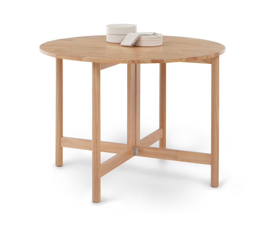 Table ronde en bois clair à quatre pieds et entretoises, surmontée de deux bols blancs empilés, d'une pile de petites assiettes blanches et d'une serviette claire pliée.