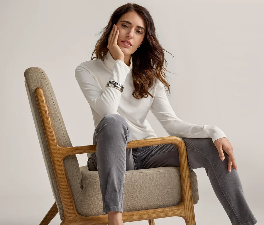 Une femme aux longs cheveux foncés et ondulés est assise sur une chaise en bois clair, vêtue d'un col roulé blanc et d'un pantalon gris. Elle pose sa main droite sur son menton et regarde devant elle. Un bracelet argenté est à son poignet gauche.