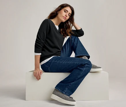 Femme assise sur un bloc blanc, portant un pull noir à col en V sur une chemise blanche, un jean bleu large et des baskets grises à plateforme.