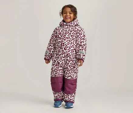 Un jeune enfant souriant se tient de face, portant une combinaison de neige blanche à imprimé léopard bordeaux. La combinaison a une capuche, des bandes réfléchissantes sur les manches et des empiècements bordeaux unis aux genoux. L'enfant porte des chaussures bleues.