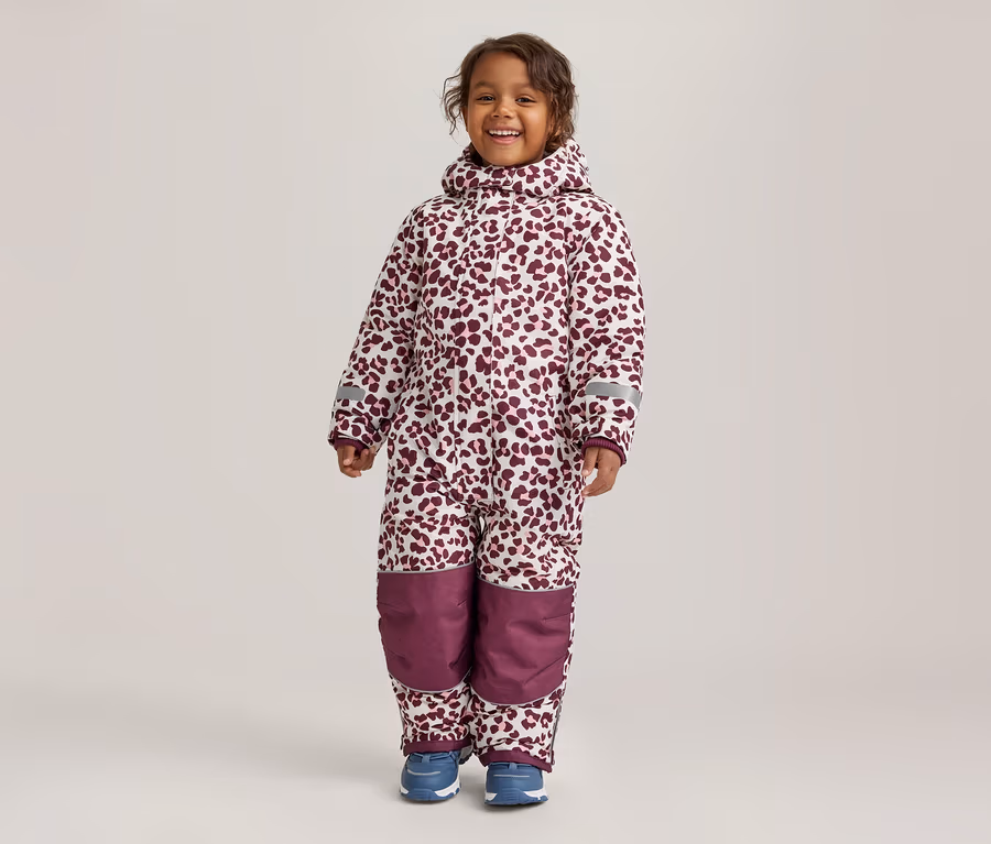 Un jeune enfant souriant se tient de face, portant une combinaison de neige blanche à imprimé léopard bordeaux. La combinaison a une capuche, des bandes réfléchissantes sur les manches et des empiècements bordeaux unis aux genoux. L'enfant porte des chaussures bleues.