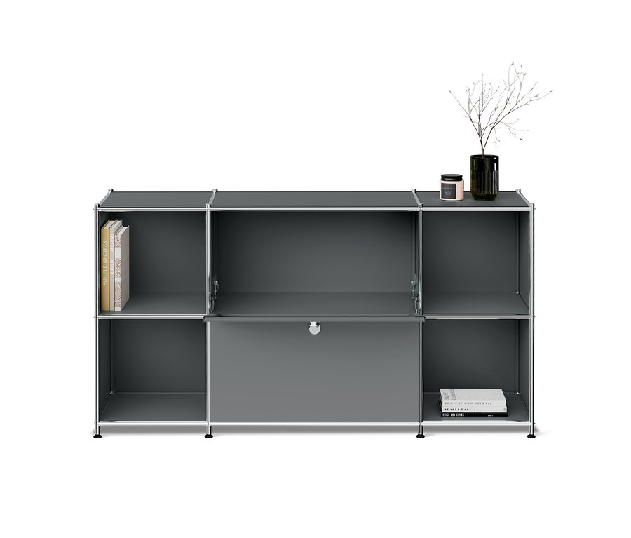 Étagère modulaire grise avec cadre chromé, six compartiments et une porte au centre en bas. Trois livres sont rangés en haut à gauche, deux livres en bas à droite. Un vase foncé avec des branches et une petite bougie sont posés sur le dessus de l'étagère.
