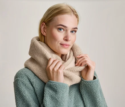 Femme blonde aux yeux bleus portant un snood en maille beige et une veste polaire vert menthe, tenant le snood des deux mains et regardant devant elle.