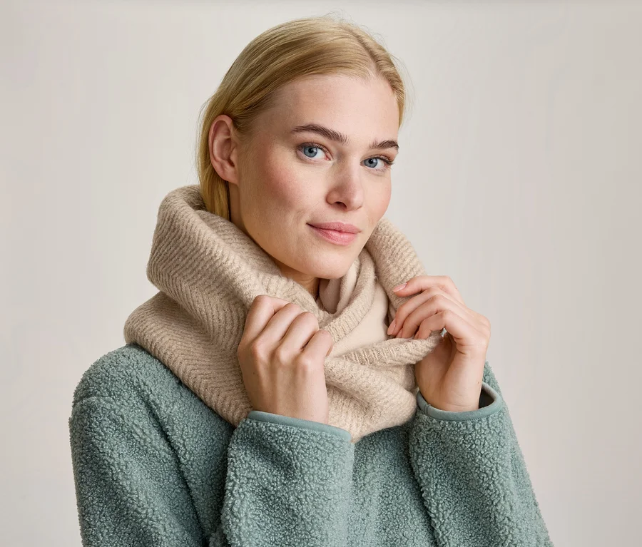 Femme blonde aux yeux bleus portant un snood en maille beige et une veste polaire vert menthe, tenant le snood des deux mains et regardant devant elle.